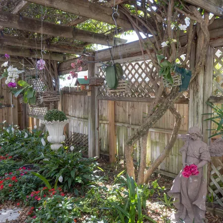 Shaded garden patio with hanging plants, colorful flowers, stone angel statue, and wooden lattice fence under pergola.