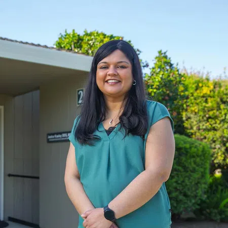 Smiling woman with long black hair in teal blouse standing outdoors near greenery and building under clear sky