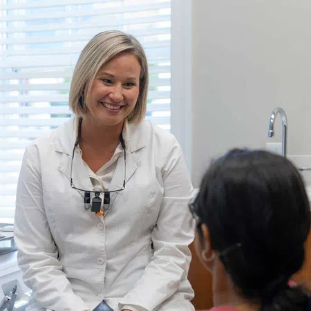 Smiling female dentist in white coat talks to patient in dental office with natural light from window.