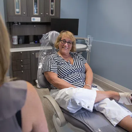 Smiling woman in casual clothes seated comfortably in a medical or dental office chair, talking to another person.
