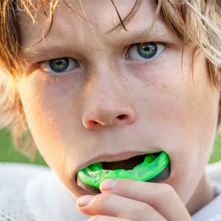 Close-up of a young athlete putting a green mouthguard in his mouth, wearing sports gear outdoors.