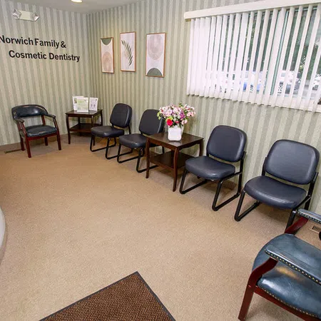 Waiting area of Norwich Family & Cosmetic Dentistry with chairs, floral arrangement, and wall decor under window blinds.