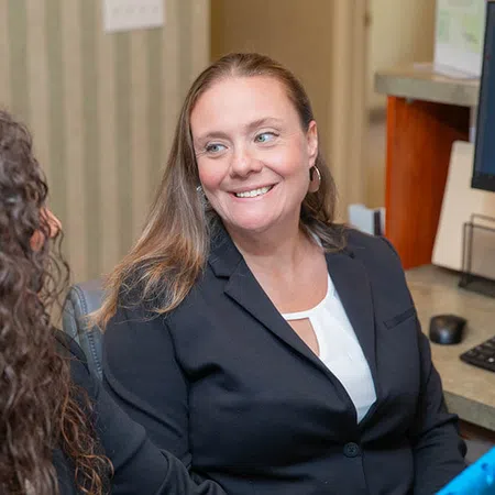 Two women in business attire smiling and talking in an office setting with a computer in the background