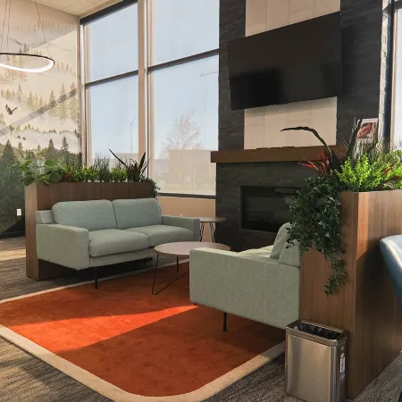 Modern office lounge with gray sofas, blue chairs, plants, a fireplace, and large windows with natural light.