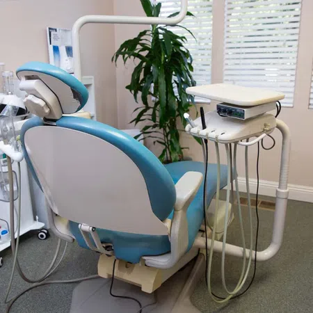 Modern blue and white dental chair with attached dental instruments and a green plant near window blinds.