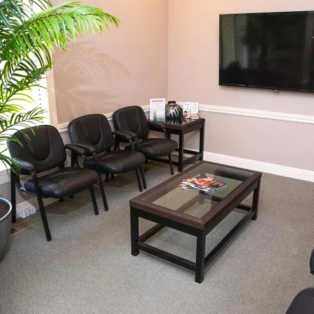 Modern waiting room with black chairs, glass coffee table, magazines, potted plant, and wall-mounted TV.