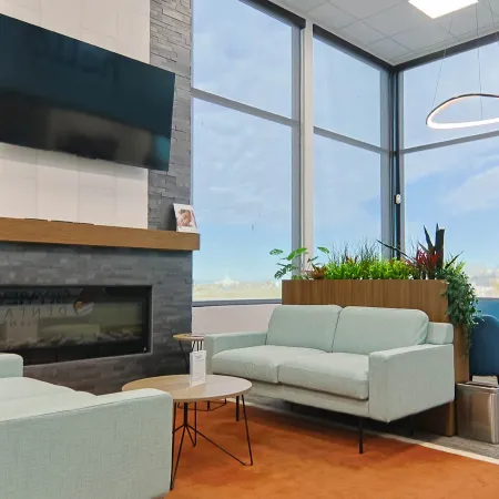 Modern office lounge with light blue sofas, large windows, indoor plants, and a mounted television above a fireplace.