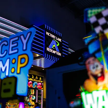 Colorful arcade game signs and plush toys under Victory Lane Redemption neon sign in a gaming center arcade.