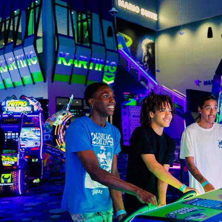 Three young men enjoying arcade games with vibrant neon lights and colorful gaming machines in the background.