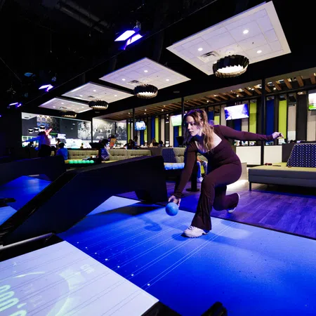 Woman playing glow-in-the-dark bowling in a modern bowling alley with vibrant blue lighting and cozy seating.