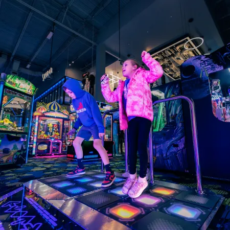 Two children playing an interactive dance arcade game with colorful lights and vibrant machines in the background.