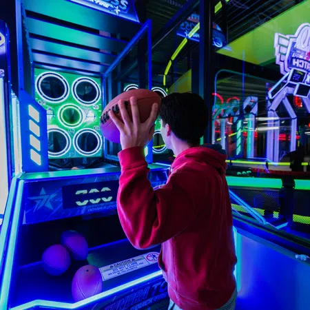 Person in a red hoodie playing a basketball arcade game in a neon-lit entertainment center.
