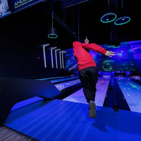 Person in a red hoodie bowling on a glowing blue lane in a modern indoor bowling alley with digital displays.