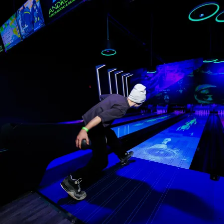 Person wearing a white beanie bowling under blacklight with glowing neon lanes and digital graphics