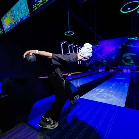 Person wearing a white beanie bowling under neon lights in a dark indoor bowling alley with digital displays.