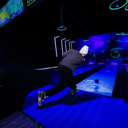 Person wearing a white beanie bowling under neon lights in a dark indoor bowling alley with glowing lanes.