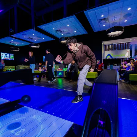 Young man bowling in a modern alley with neon blue lighting and spectators watching from couches.