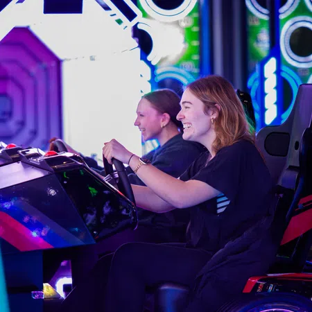 Two women enjoying a colorful arcade racing game in a vibrant gaming center with neon lights.