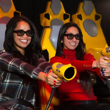 Two women wearing 3D glasses, holding yellow laser guns in a colorful arcade setting.