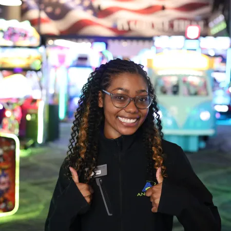 Smiling arcade employee giving thumbs up in a colorful game arcade with bright lights and machines.