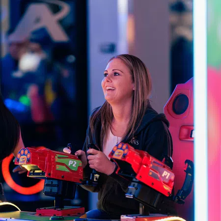 Smiling woman playing a red arcade shooting game with dual guns in a vibrant gaming environment.