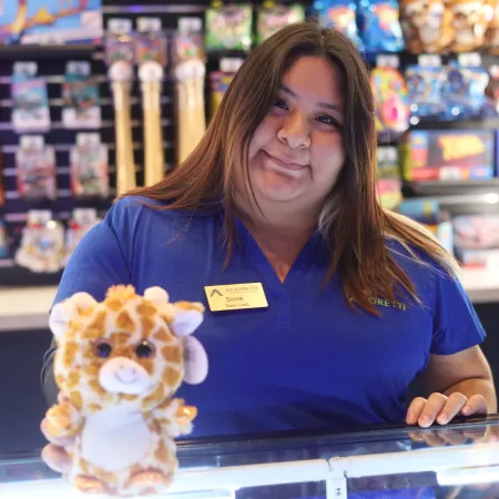 A smiling employee poses at a toy store, holding a stuffed giraffe toy, colorful merchandise in the background.
