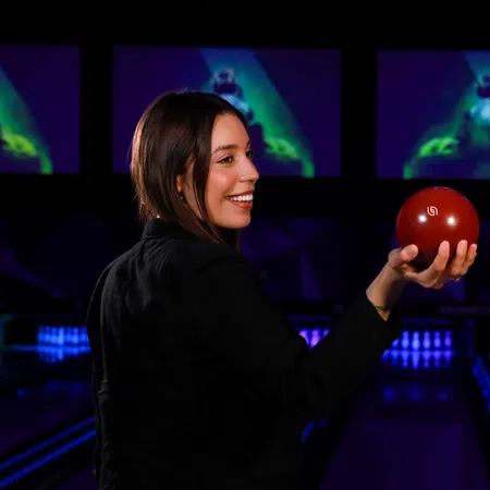 Smiling woman holding a red bowling ball in a vibrant bowling alley with screens illuminated in the background.