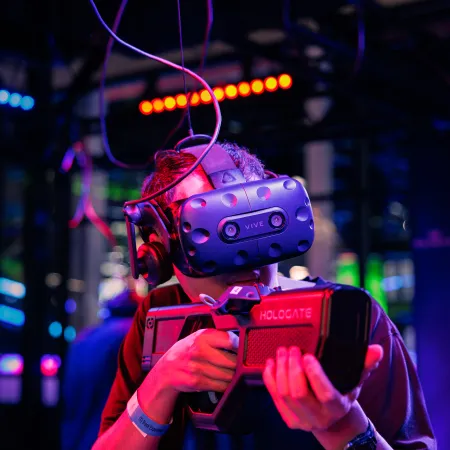 Person wearing HTC Vive VR headset and holding HOLOGATE VR gun in a colorful gaming arcade environment.
