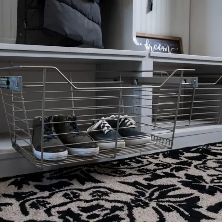Modern entryway storage with wire basket holding shoes under white seating bench on patterned rug.