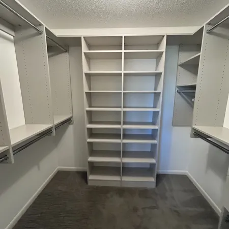 Empty white walk-in closet with multiple shelves, hanging rods, and grey carpeted floor under bright light