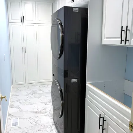 Modern laundry room with black stacked washer and dryer, white cabinets, and marble tile flooring under bright lighting.