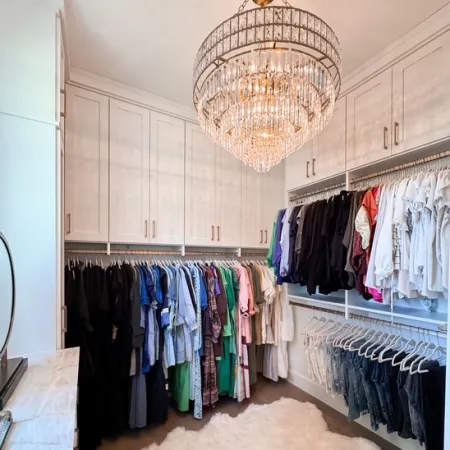 Spacious walk-in closet with organized hanging clothes, white cabinetry, a crystal chandelier, and a plush rug.