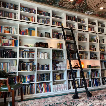 Large white bookshelf filled with books, a rolling ladder, green leather chair, desk, and decorative ceiling mural.