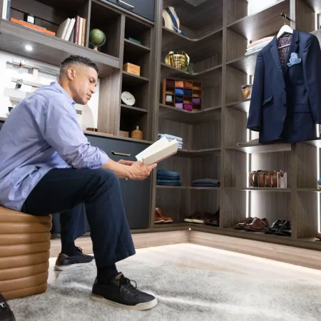 Man sitting on a leather ottoman reading a book in a modern walk-in closet with shelves and clothing.