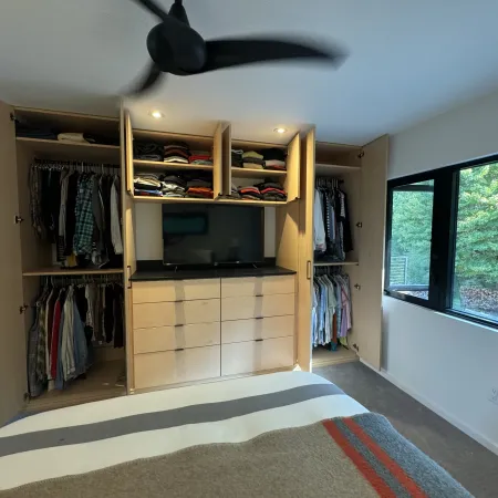 Modern bedroom closet with open wooden cabinets, folded clothes, hanging shirts, and a view of green trees through a large window