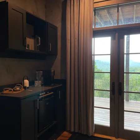 Modern kitchenette with black cabinets, coffee maker, and large window with beige curtains overlooking green landscape.