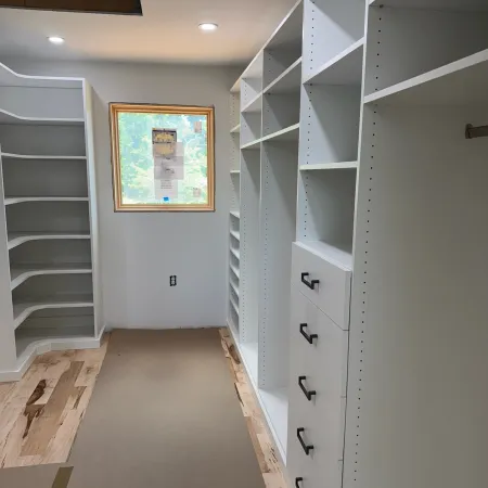 Empty white walk-in closet with built-in shelves, drawers, and a wooden floor under construction with a window.