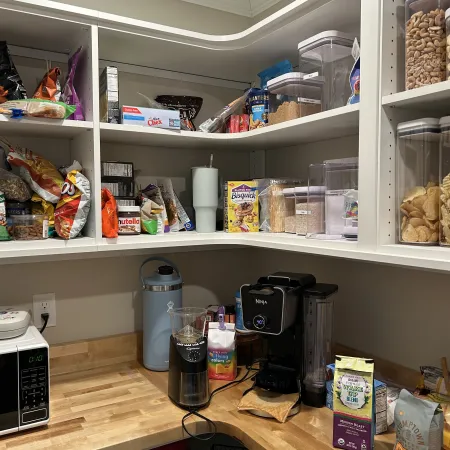 Organized pantry shelves with snacks, cereal, jars, blender, coffee grinder, microwave, and wooden countertops in kitchen corner.