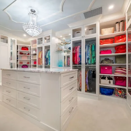 Luxurious walk-in closet with white cabinetry, glass doors, marble island, and crystal chandelier lighting