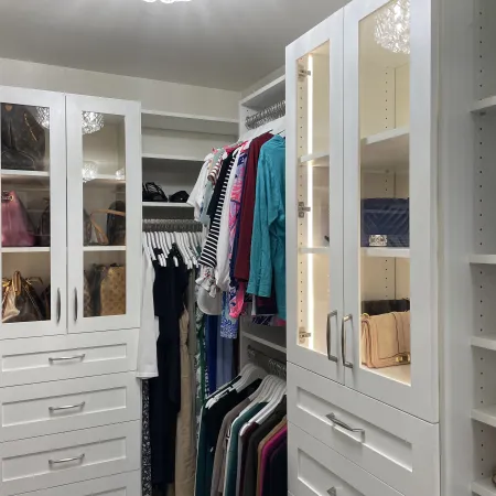 Organized walk-in closet with white shelves, drawers, hanging clothes, handbags, and shoes.