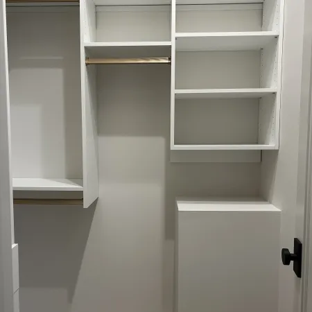 Empty modern white closet with shelves, hanging rods, and a pedestal in a small room with beige carpet.
