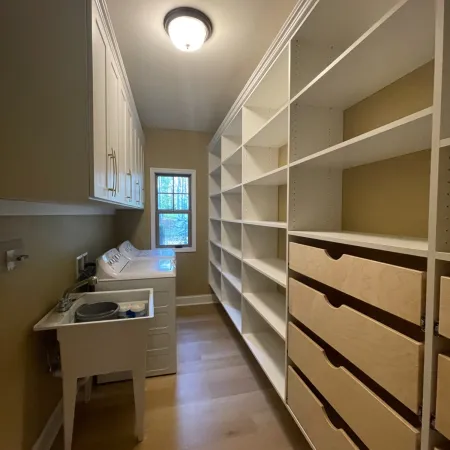 Laundry room with white cabinets, open shelving, washer, dryer, and utility sink under a window.