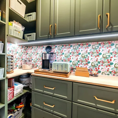 Organized pantry with green cabinets, floral backsplash, toaster, coffee maker, and various storage baskets and jars.