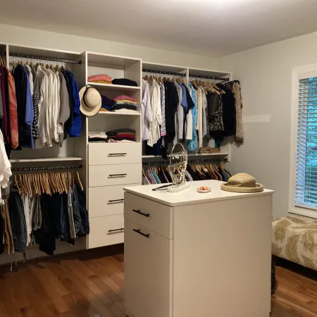 Spacious walk-in closet with organized hanging clothes, central dresser island, window, and cushioned bench seating.
