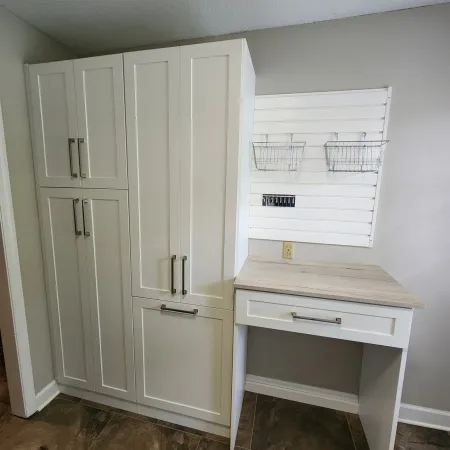 White storage cabinets and a desk with a wood countertop next to a wall organizer with wire baskets.