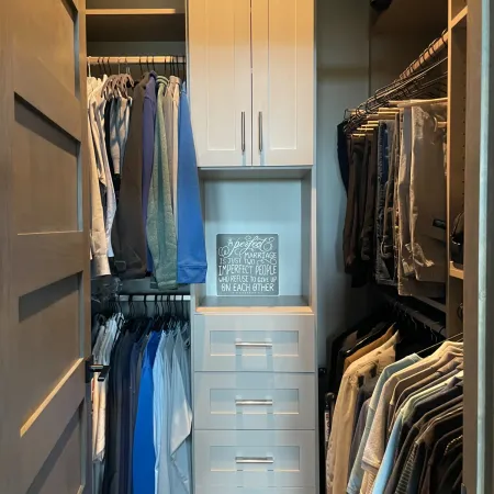 Organized walk-in closet with hanging pants and shirts, white cabinets, and a framed inspirational quote on the wall.