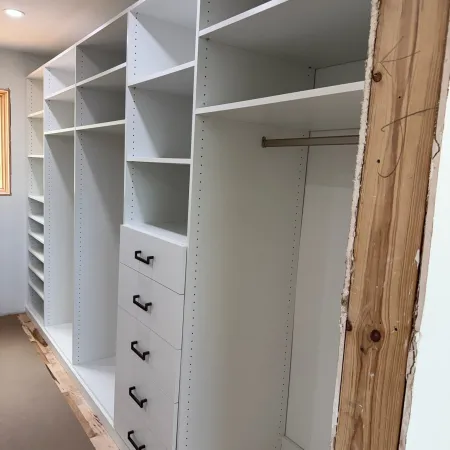 Custom white built-in closet with open shelves, drawers, and hanging rod under construction in a room.