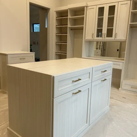Light wood walk-in closet with central island, drawers, shelves, and built-in cabinets in a bright room.