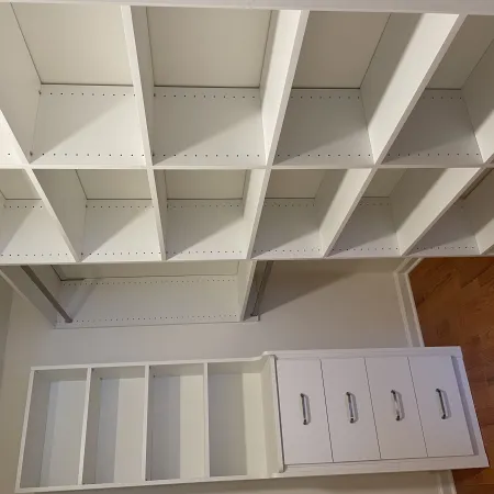 Empty white closet organizer with shelves, drawers, and hanging rods on wooden floor in a room.