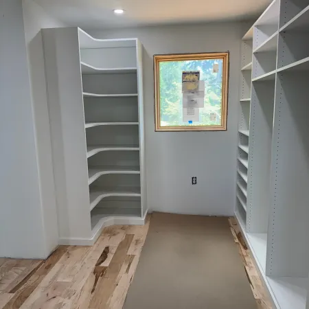 Bright walk-in closet with white built-in shelves and hardwood floor under construction with window light.